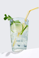 Mojito cocktail with ice cubes is contained in highball glass with a straw, lime slices and a sprig of mint and isolated on a table edge. The showy illustrative picture is made on the white backdrop.