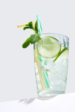 Mojito cocktail with ice cubes is contained in highball glass with straws, a lime slice and a sprig of mint and isolated on a table edge. The showy illustrative picture is made on the white backdrop.