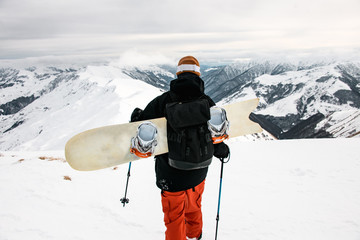 Staying snowboarder with ski equipment on background of snow-capped mountains and cloudy sky.