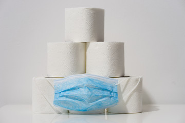Front view on white toilet paper rolls stacked on the table in front of the wall background with blue protective mask during the virus epidemic situation concept