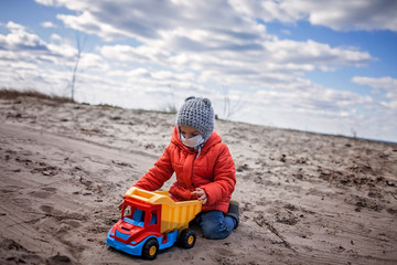 An adorable 4-5 years old boy in red coat wearing respirator mask playing with toy cargo car alone