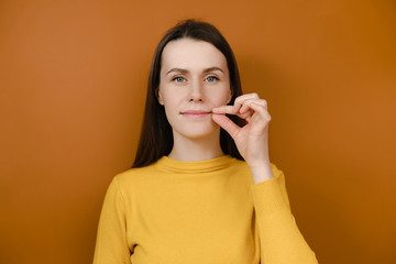 Mysterious smiling woman looks mysteriously, aside zips her mouth shut, promises to keep secret, holds fingers near cheek, hides information, dressed in yellow sweater, isolated on orange background