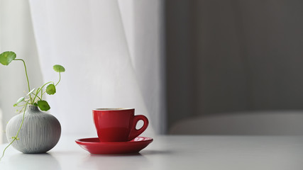 ivy in vase and red ceramic coffee cup with coasters putting on the white modern desk over living room curtain as background.