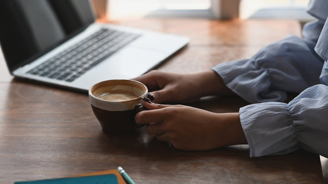 Cropped image of beautiful woman's hands holding a ceramic coffee cup in hands while sitting at the wooden working table in front her computer laptop.