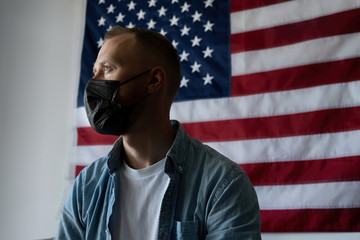 Obraz premium Man in a black medical mask sits on a home quarantine against the backdrop of the American flag. Self isolation due to coronavirus. Measures to curb the spread of Covid-19. The health of the country.