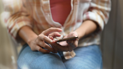 Cropped image of beautiful woman using/holding smartphone in her hands while sitting at the working desk over modern living room as background.