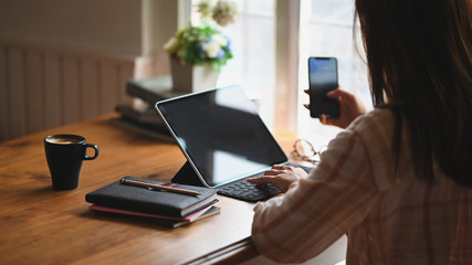 Cropped image of secretary woman's hand holding a smartphone in her hand while sitting and using a black blank screen computer tablet with keyboard case at the wooden working table in modern office.