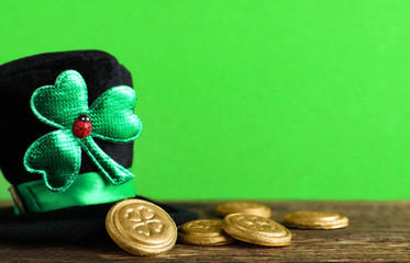 Black leprechaun hat and gold coins on wooden table against green background, space for text. St....