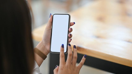 Cropped image of beautiful woman holding a white blank screen smartphone in hands while sitting at the wooden working desk over the modern office as background.