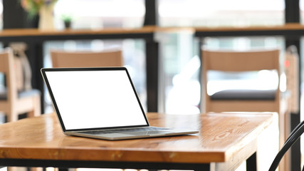 Photo of white blank screen computer laptop putting on wooden working desk over modern cafe/restaurant as background. Orderly workplace concept. Space for advertisement.