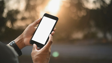 Cropped image of smart man's hands holding a cropped black smartphone with white blank screen over blurred sunny outdoors as background.