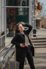 Portrait of a girl with a drink in her hands, and her eyes closed, enjoys the sun's rays, vertical photo. Young woman on a city street with coffee in her hands on a sunny day.