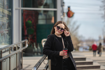 Fototapeta premium cheerful woman on a city street drinks coffee on a sunny day and looks at the camera. coffee to go. Portrait of a girl with a drink in hands