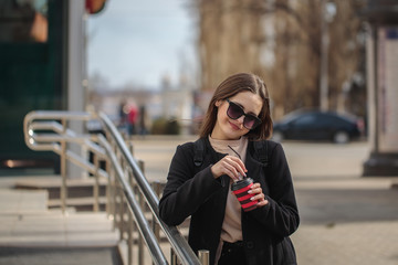 young woman on a city street drinks coffee in a sunny day in sunglasses and looks away. coffee to go. Portrait of a girl with a drink in hands