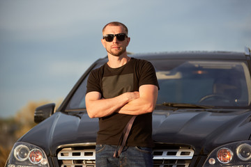 Portrait of a successful self-confident man standing in front of his nice black car with crossed hands, wearing dark sunglasses, black t-shirt and jeans, close-up