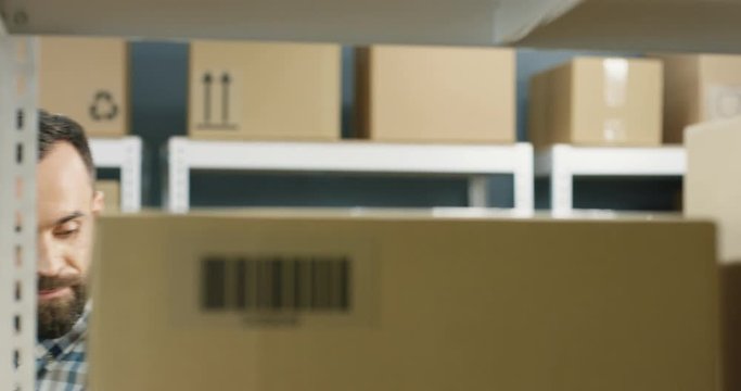 Good looking Caucasian young mailman putting parcel with bar code on shelf in post office store. Close up of carton box in mail storage. Courier bringing cardboard box to postal room.