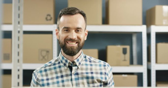 Close up of Caucasian good looking postman taking carton box with bar code from shelf and smiling to camera in post office store. Portrait of man postal worker with happy smile and parcel in hands.