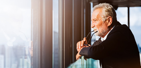 Retirement concept. Senior grey-haired businessman standing and looking to right hand at modern business lounge high up in an office tower.