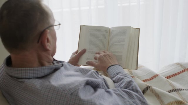Retired man reading a book in the bed. Slow motion shot, man turning the page