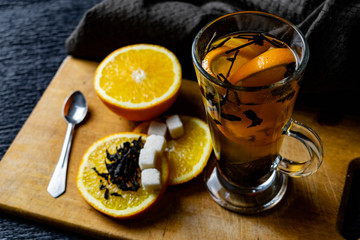 Citrus drink, tea with petals in a transparent glass mug with slices of sweet orange and refined sugar. Slicing fruit on a wooden Board with a brown napkin in a cozy dark background in a low key