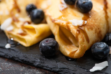 close up view of tasty crepes with blueberries, mint and coconut flakes served on board on textured background