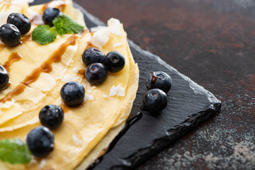 close up view of tasty crepes with blueberries, mint and coconut flakes served on board on textured background