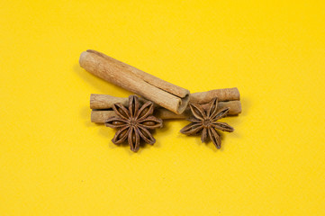 two sticks of cinnamon, one lying on top of the other, two stars of star anise, close-up, on a yellow background side view