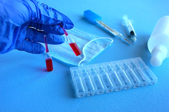 A hand in a blue medical glove holds ampoules with red liquid, next to a syringe, medicine, chlorhexidine, antispetic and thermometer. - Powered by Adobe