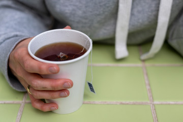 The girl holds in her hand a disposable white cup with black tea. Tea bag inside. Popular takeaway drinks concept. A cup on a green table. Horizontal view.