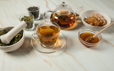 Healthy and delicious herbal tea with honey. A teapot, tea in a glass cup, sweet golden honey and a set of dried herbs in a porcelain mortar with a pestle close-up on a pastel green marble background.