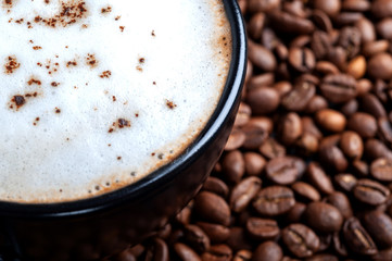  A black cup of black coffee with white foam close-up, stands on a table where coffee beans are scattered. top shot copyspace