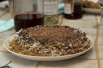 Grandma’s homemade cake sprinkled with grated chocolate. A cake stands on a rustic table covered with a tablecloth, in the background a bottle of red and white wine is out of focus.