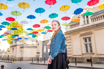 Beautiful girl walking under umbrellas in the city. Cute teenage girl in black dress posing against a background of multicolored umbrellas alley. Spring sunny concept. 8 March. Happy Women's Day.