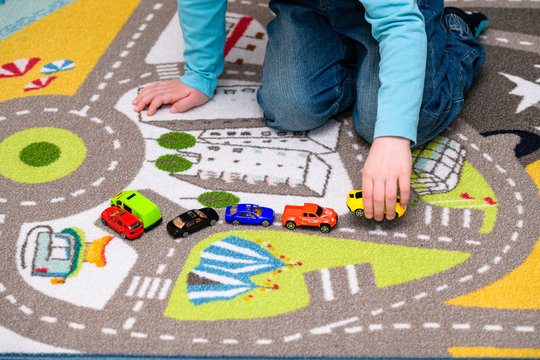 Five year old boy playing and lining up toy cars on a playing mat with roads. The cars have vivid colors and the boy is dressed in blue jeans and a blue shirt. He is holding a yellow toy car.