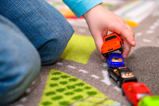 Close-up of five year old boy playing and lining up toy cars on a playing mat with roads. The cars have vivid colors and the boy is dressed in blue jeans and a blue shirt. He is holding a red pickup t