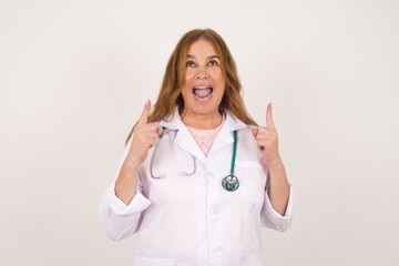 Charming doctor woman with positive expression, points up with both index fingers, wearing medical...
