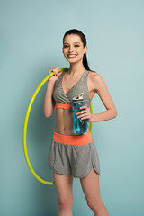 cheerful athletic woman holding hula hoop and sports bottle with water on blue