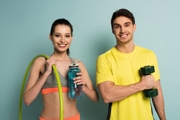 happy athletic couple holding dumbbell, hula hoop and bottle of water on blue