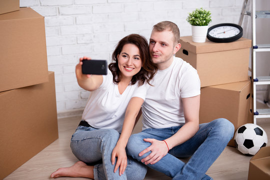 moving day - portrait of young couple taking selfie photo with cardboard boxes in their new house or flat