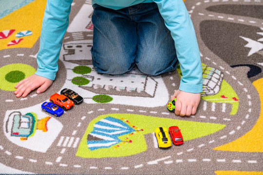 Five year old boy playing and lining up toy cars on a playing mat with roads. The cars have vivid colors and the boy is dressed in blue jeans and a blue shirt. He is holding a toy car.