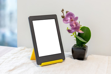 Modern gray electronic book or tablet with a blank mockup screen on a yellow stand next to an artificial pink orchid on the kitchen table with a linen tablecloth by the window