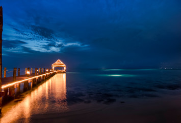 Wooden pier illuminated at night with blue sky background, and reflections on the Caribbean sea in the island of Cozumel Mexico