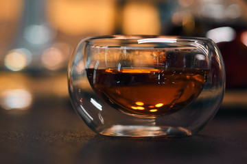 Transparent bowl with black tea on a wooden table. Blurred background of bright lights. Horizontal picture.
