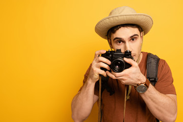 Obraz premium handsome tourist in hat with backpack taking photo on camera on yellow