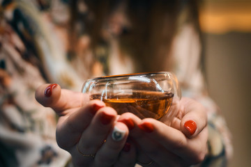 Hands of a girl holding a transparent bowl with black tea. Bright highlights on a glass. Cozy picture.