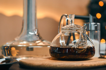 A transparent teapot of black tea on a wooden stand. In the background is a hookah and sugar.
