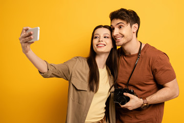 couple of happy tourists with photo camera taking selfie on smartphone on yellow
