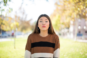 Pleasant looking young female, keeps lips as going to kiss someone, has glad expression, grimace face. Standing outdoors. Beauty concept.