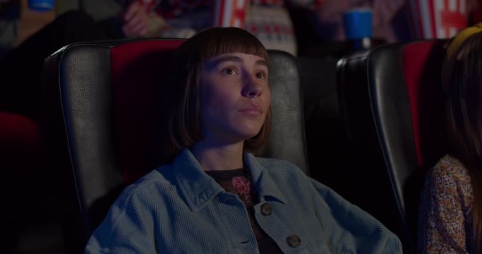 Front view of millennial girl enjoing fascinating film in movie theatre. Hipster gil with nose ring chewing popcorn and watching movie in cinema. Concept of leisure and entertainment.
