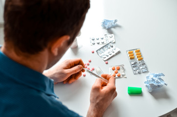 A young man in blue shirt holding a thermometer and many pills on a table at home